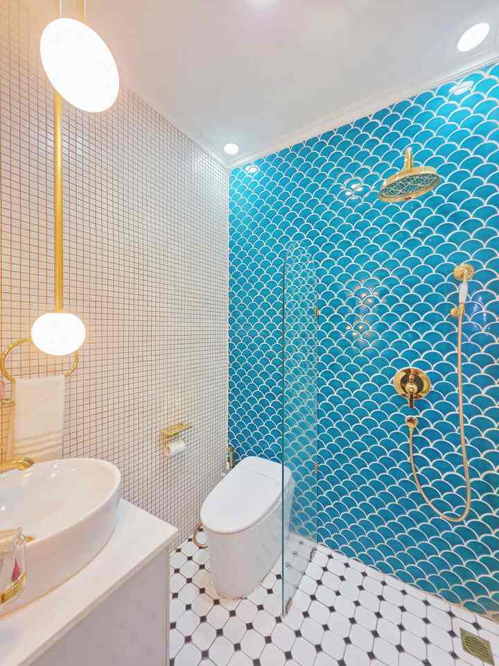 A narrow modern bathroom featuring white and blue mosaic tiled walls, gold shower fixtures, and elegant lighting