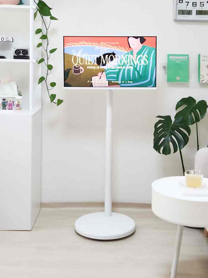 Bright white and natural toned living room featuring a central moving TV stand with monitor, plants, and clean decor for a cozy atmosphere