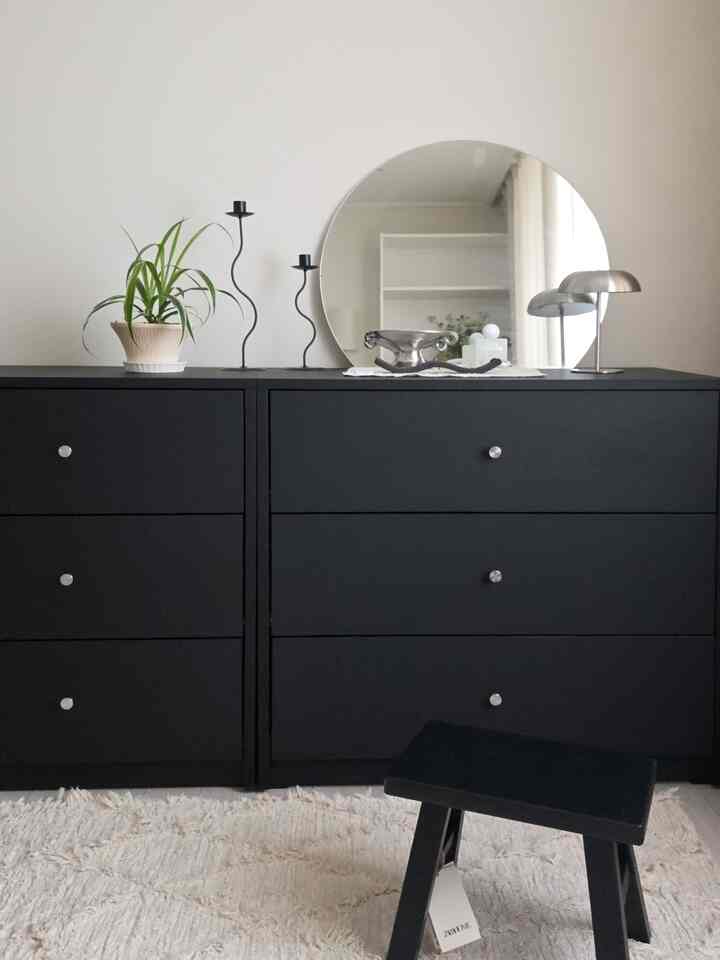 黒いチェストが中央に配置された寝室空間、植物と丸い鏡、小さな黒いスツールが配されたモダンで清潔感のあるインテリア