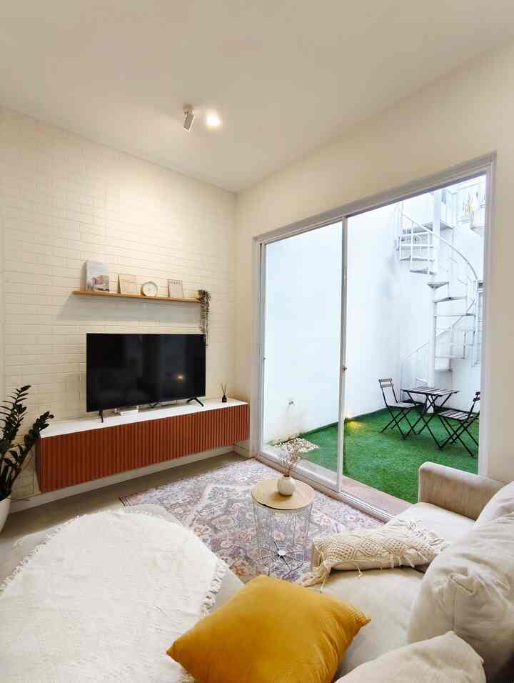 White and natural toned living room featuring beige sofa, plants, and a balcony visible through sliding glass door, creating a cozy atmosphere
