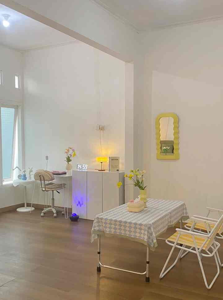 Bright white-walled dining room with brown wood-tone flooring, featuring a checkered tablecloth, yellow framed mirror, and glowing table lamp creating a cozy atmosphere