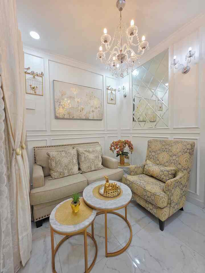 White and gold toned classic living room featuring beige sofa, patterned armchair, and crystal chandelier with elegant ambiance
