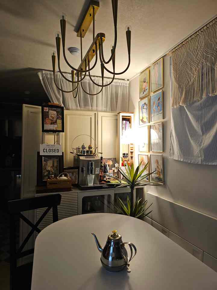 白とゴールドのトーンが調和するリビング空間、丸いダイニングテーブルと多くのゴールドフレームの壁アート、コーヒーマシンが配置された雰囲気のあるおうちカフェ