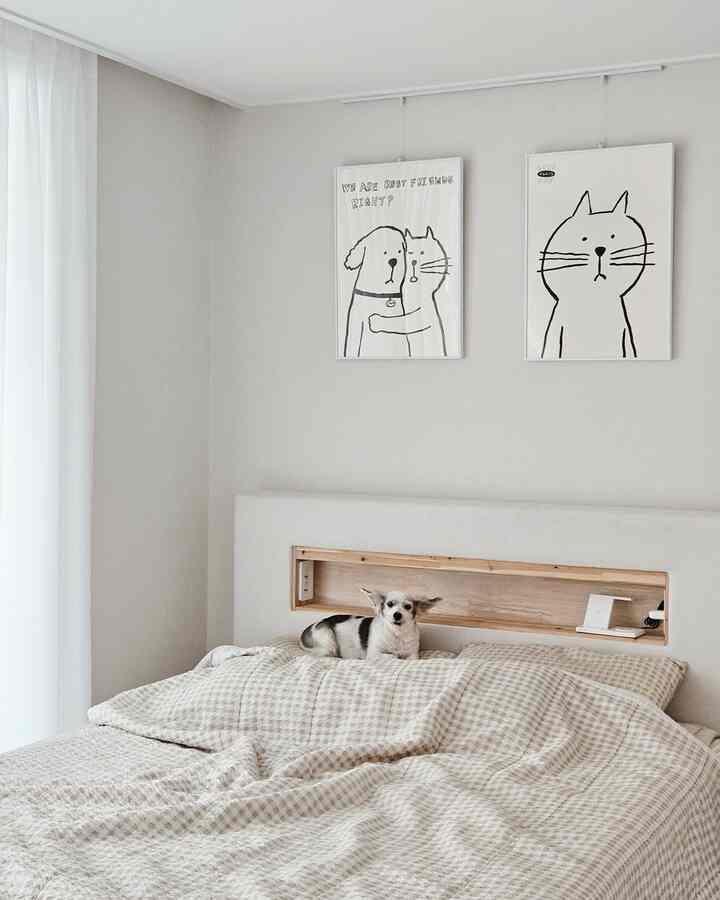 白い壁とベージュのチェック柄の寝具のシンプルな寝室、ベッドの上に犬がいて、壁には猫と犬のイラストが飾られている印象的な空間