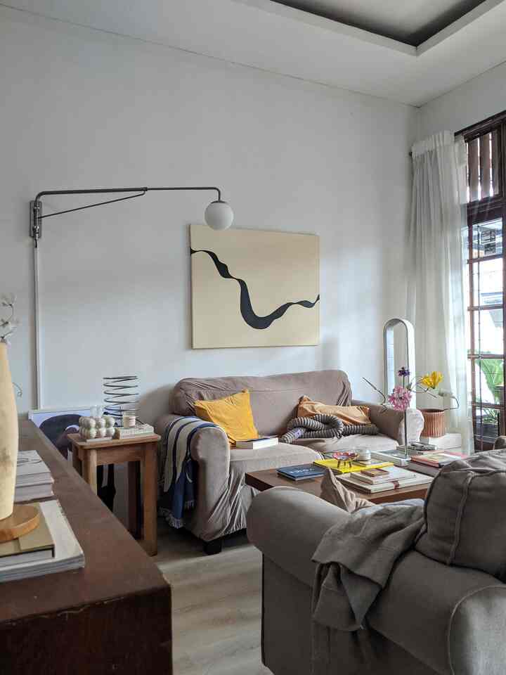 A white-walled living room with brown wooden furniture, featuring sofas and a coffee table stacked with books creating a cozy atmosphere
