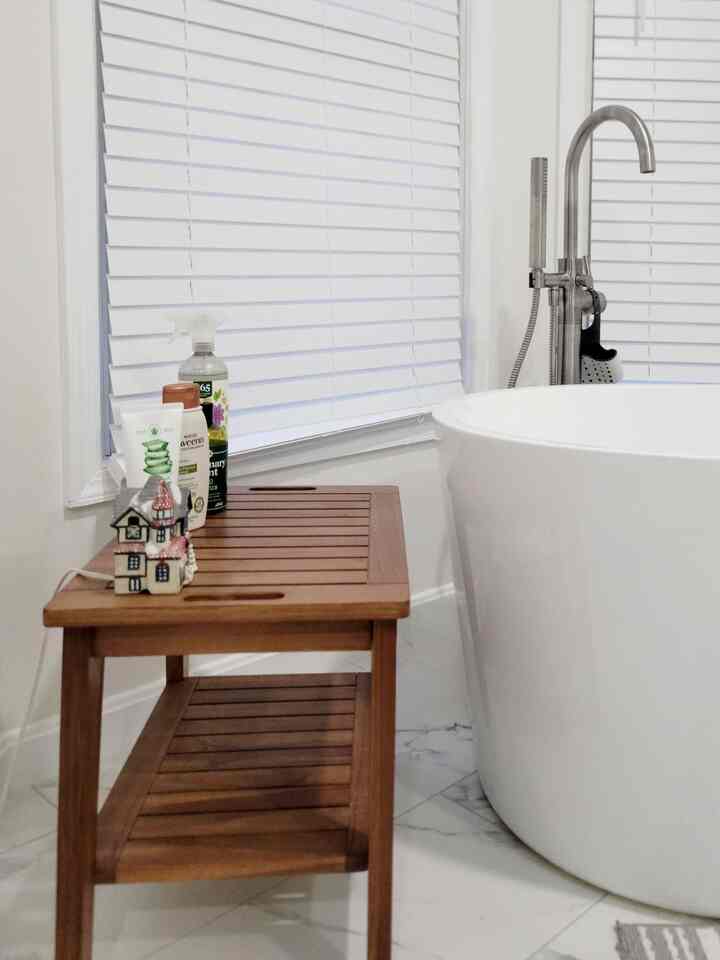 A clean bathroom in white and wood tones featuring a freestanding bathtub next to a practical wooden bench