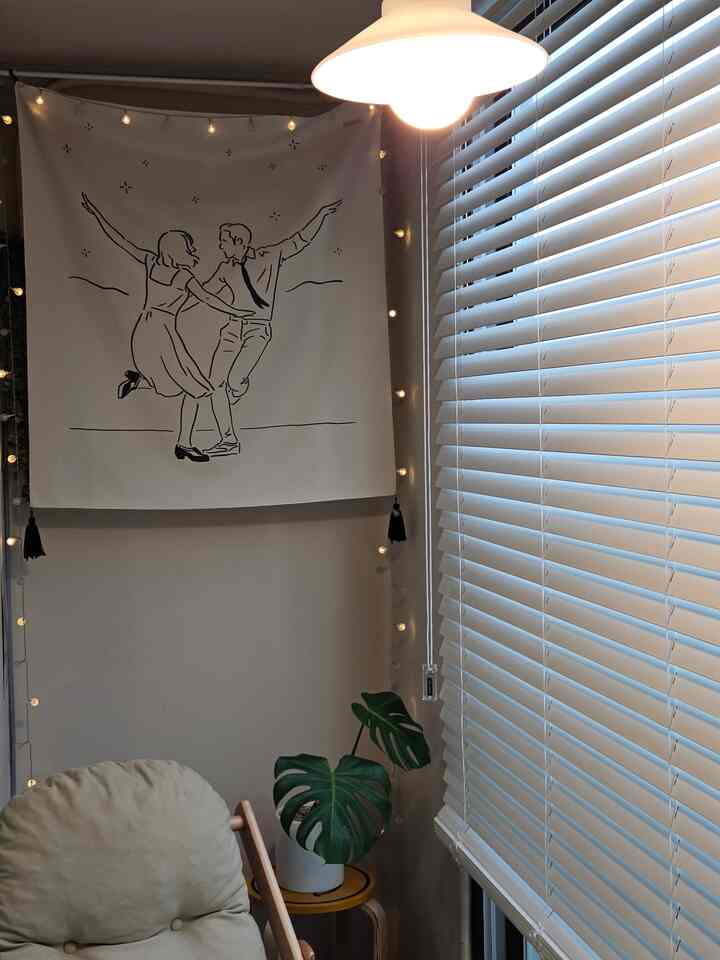 ベージュのアームチェアとモンステラの植物、白いブラインドがあるナチュラルなリビングコーナー、壁に踊るカップルの絵画が飾られている雰囲気のあるインテリア