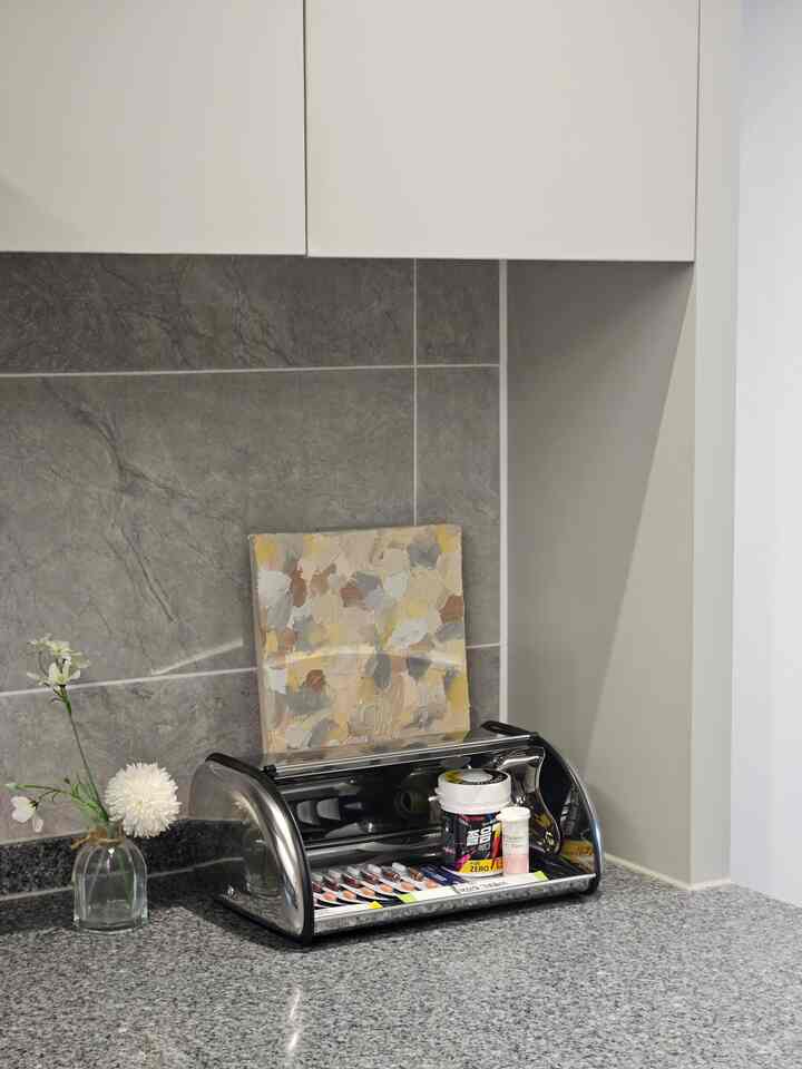 Gray marble tiled wall and gray granite countertop featuring a stainless steel roll-up bread box and small flower vase in a neat kitchen space