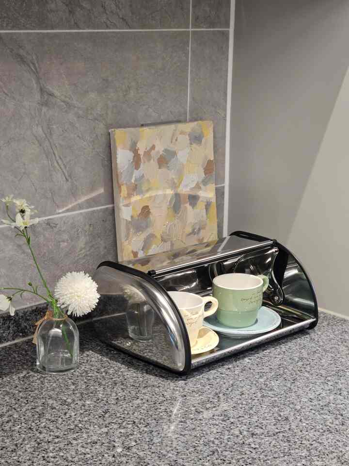 Gray marble countertop featuring a stainless steel bread box with two mugs, beige abstract painting behind, and a clear flower vase, creating a neat kitchen corner