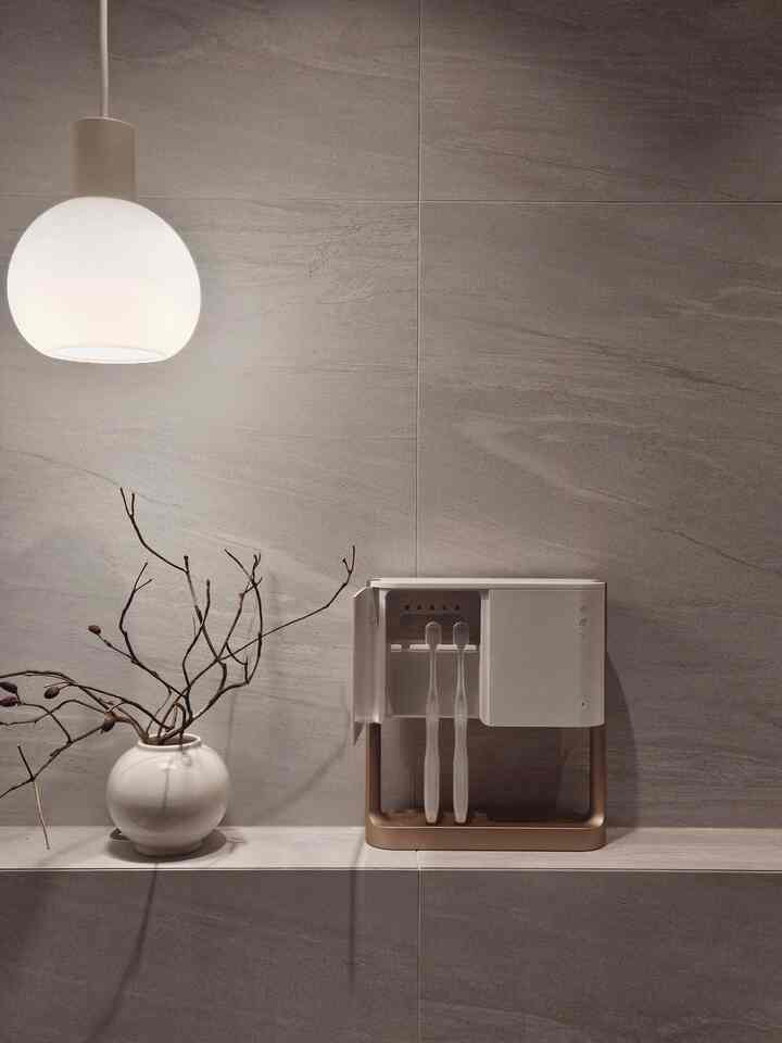 グレーのタイル壁に白いペンダントライト、丸い花瓶と歯ブラシ殺菌器が配置されたシンプルなバスルームのインテリア