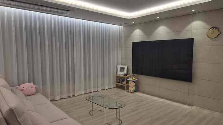 ベージュとナチュラルカラーのリビング、透明な強化ガラスの楕円形テーブル、ベージュのソファ、壁掛けテレビ、マンガキャラクターの壁時計が配置された雰囲気のあるインテリア