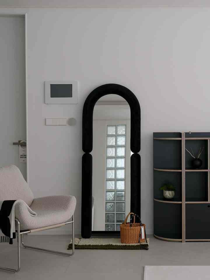白い壁と床のモダンなリビング、中央に黒いベルベットフレームの全身鏡、左にアームチェア、右に収納棚が配置された印象的な空間