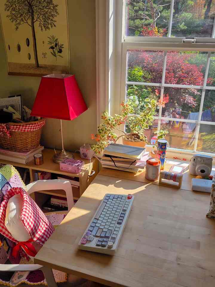 ベージュ色の壁と大きな窓のあるホームオフィススペース、木製の机と赤いテーブルランプ、植物が配置された温かみのあるインテリア