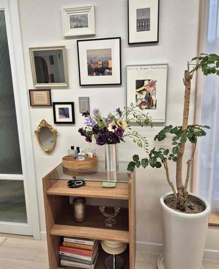 ナチュラルトーンの小さなコーナースペース、ガラス天板の木製チェスト、多数の額縁と鏡、紫色の花が生けられた花瓶、大きな観葉植物が配置された雰囲気のあるインテリア