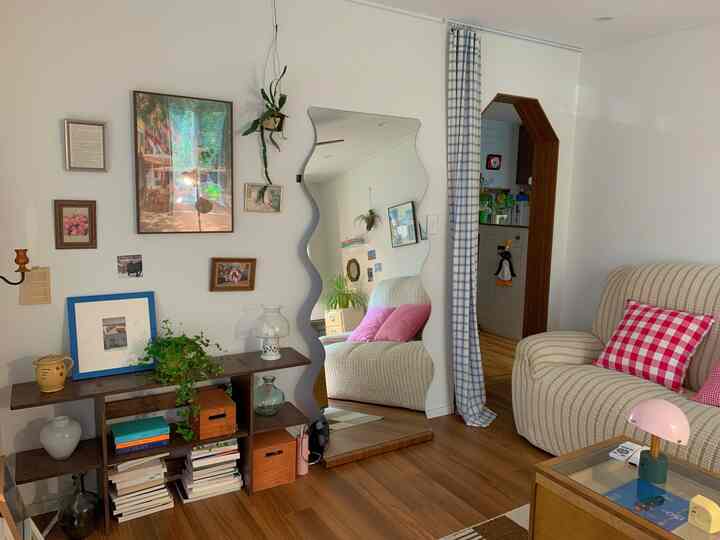 白い壁と木目調の床が調和するリビング、波形の全身鏡と額縁の壁飾り、チェック柄クッションが映えるシンプルで自然な雰囲気のインテリア