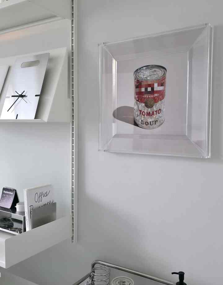 Minimal white-toned wall space featuring a clear framed artwork, aluminum clock, and a desk with organized office items