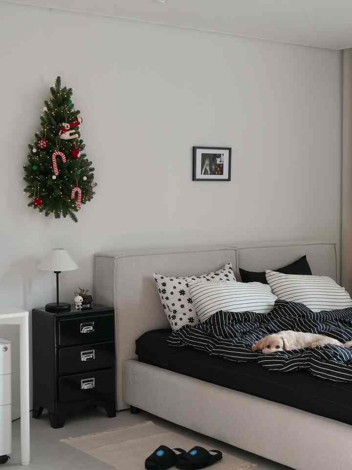 黒と白のトーンが調和する寝室、ファブリックベッドとブラックのナイトテーブル、壁に掛けられたクリスマスツリーが配置された雰囲気のあるインテリア