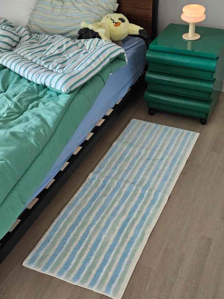 Bedroom in green and blue tones featuring a bed, green nightstand, and pastel striped bath mat on wooden floor creating a cozy atmosphere