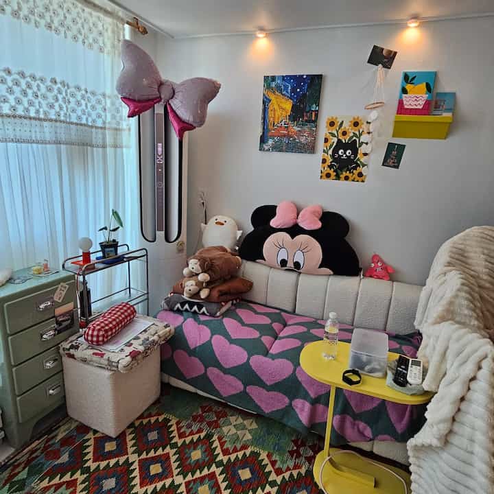 白い壁とピンクのハート柄ブランケットを配したミッドセンチュリーモダンのリビング、カラフルなおもちゃとソファが置かれた雰囲気のある空間