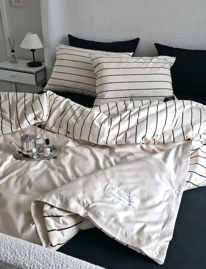 ナチュラルカラーとストライプ柄の寝室、白いナイトテーブルとシルバーティーポットが配置されたモダンな印象の空間