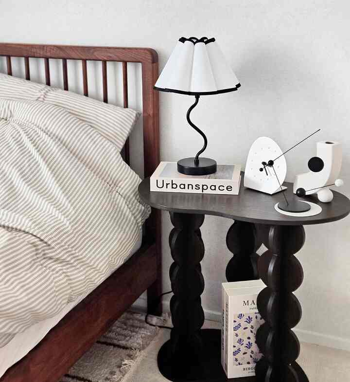 Natural wood frame bed with black and white nightstand featuring table lamp, minimal bedroom interior