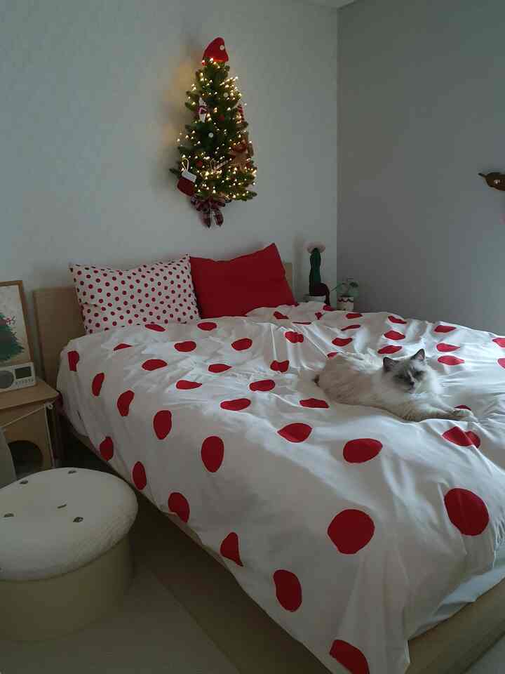 ホワイトとレッドのドット柄寝具がある居心地の良い寝室、壁に飾られたクリスマスツリーとベッドにいる猫のいる雰囲気のあるインテリア