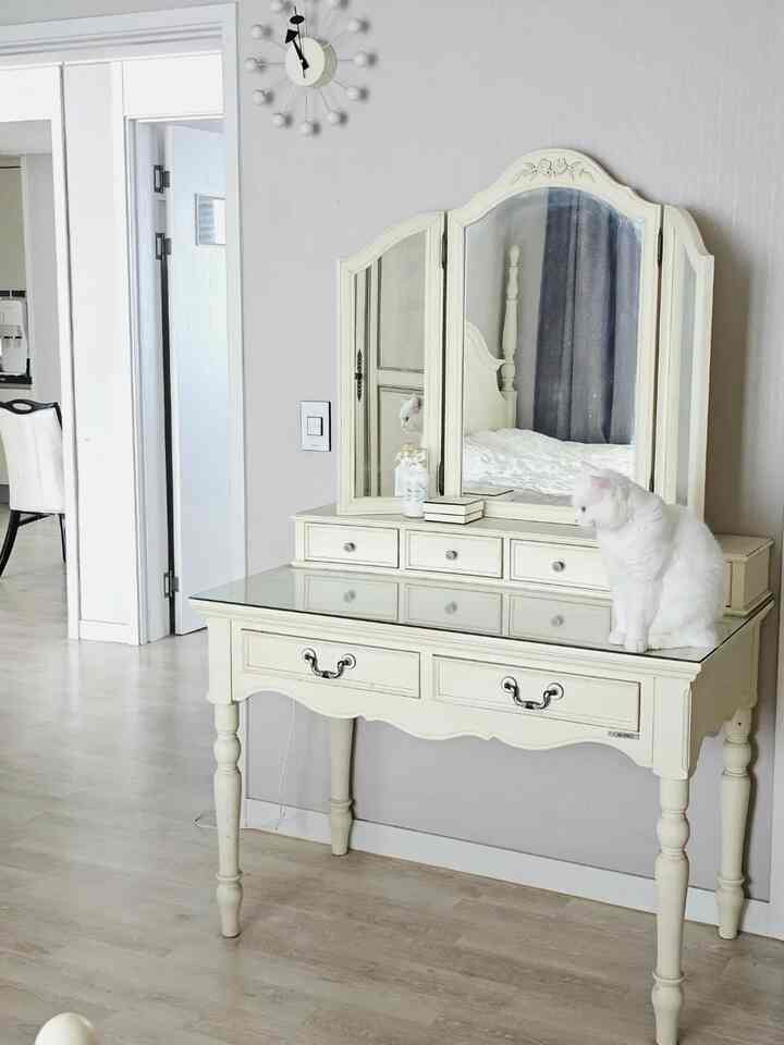 白とナチュラルカラーの寝室、アンティーク風の化粧台と鏡、そして上に座る白い猫が配置された落ち着いた雰囲気のインテリア