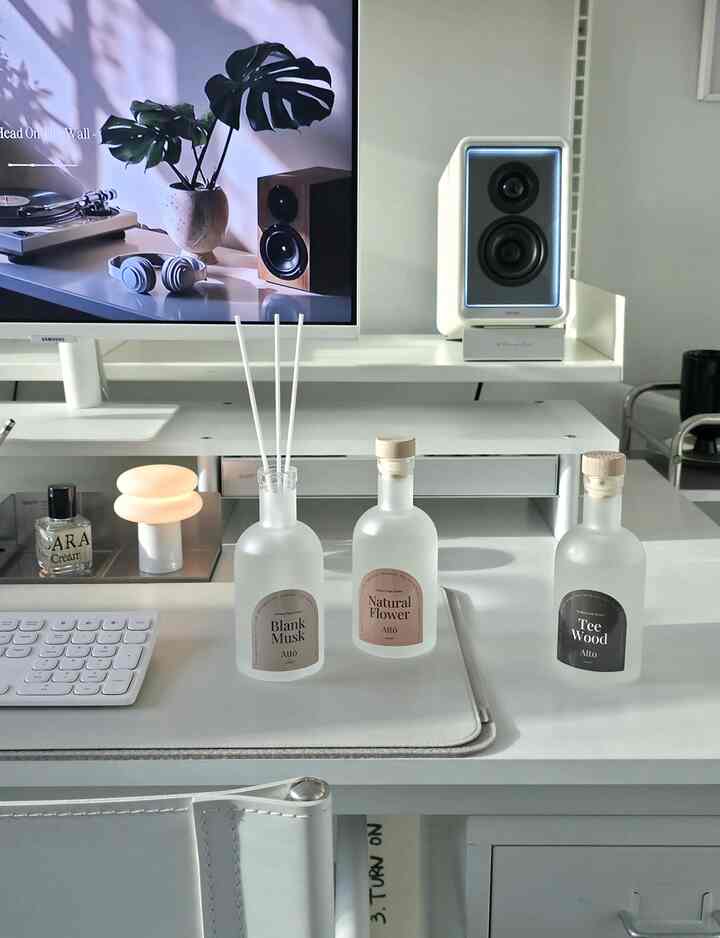 White-toned home office space featuring a desk with diffusers, monitor, and speaker arranged neatly