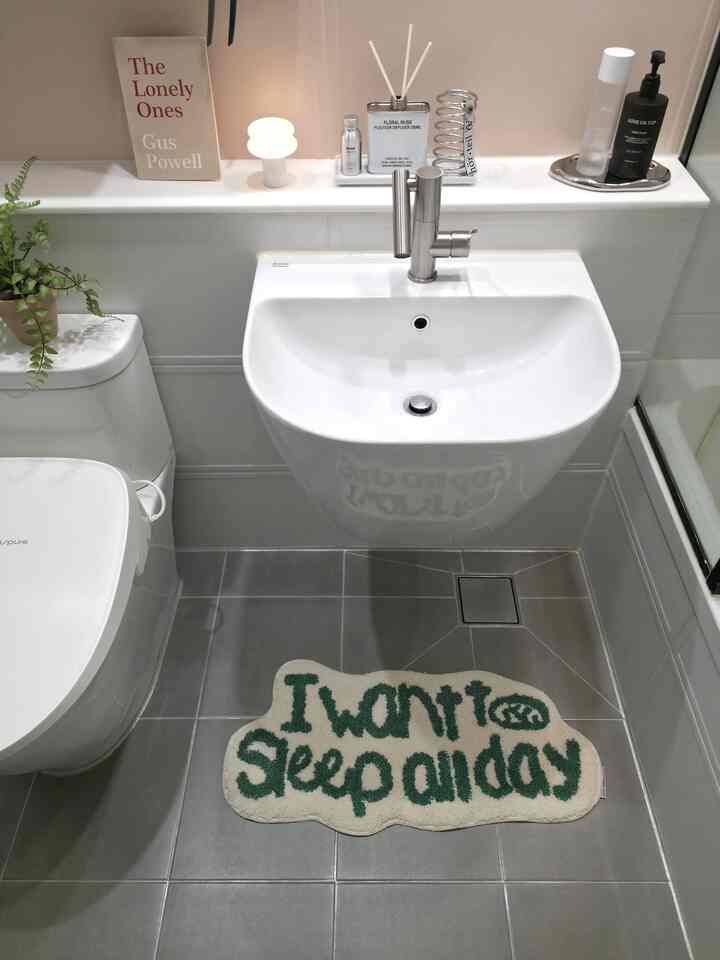 グレーのタイル床と白い洗面台およびトイレがある清潔なバスルーム、床には「I want to sleep all day」と書かれたラグが敷かれている