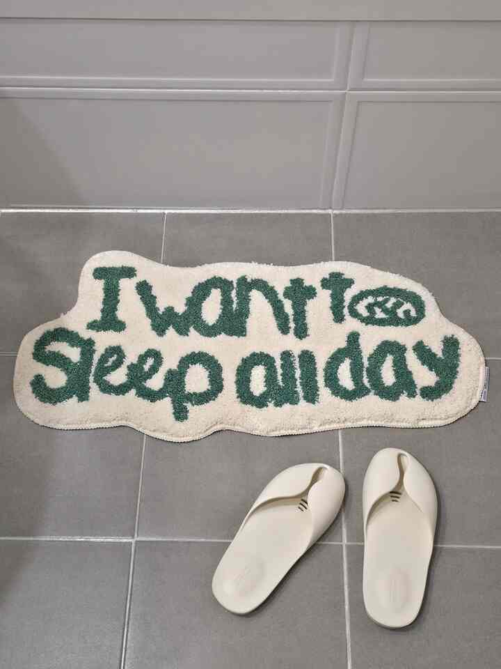 グレーの床タイルと白い壁タイルの上に「I want to sleep all day」と書かれたミニラグが配置されたバスマットのある空間