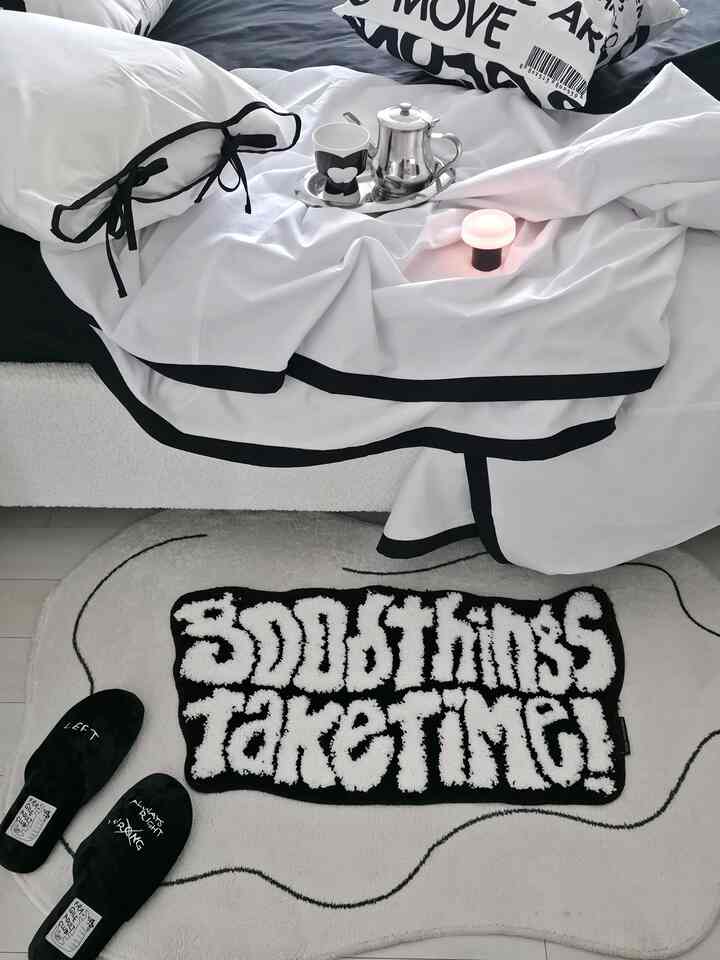 黒と白のトーンの寝室、中央にユニークなレタリングのバスマットと白い寝具が配置されたモダンな空間