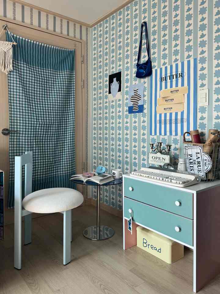 パステルブルーとホワイトのトーンで彩られた狭いホームオフィス空間、ブルーのパターン壁紙とミニデスク、サイドテーブル、アークダイニングチェアが配置された雰囲気のあるインテリア