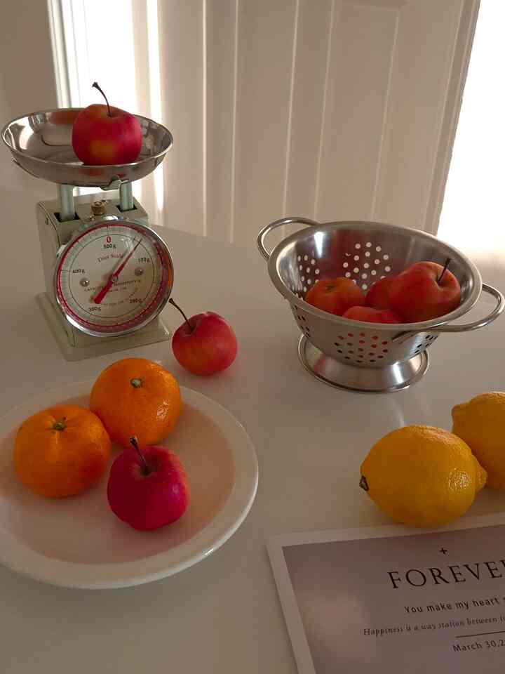 柔らかなホワイトトーンのキッチンテーブルに、ダルトンのキッチンスケールと皿、ステンレス製のフルーツざるが配置された、温かみのあるキッチンのインテリア