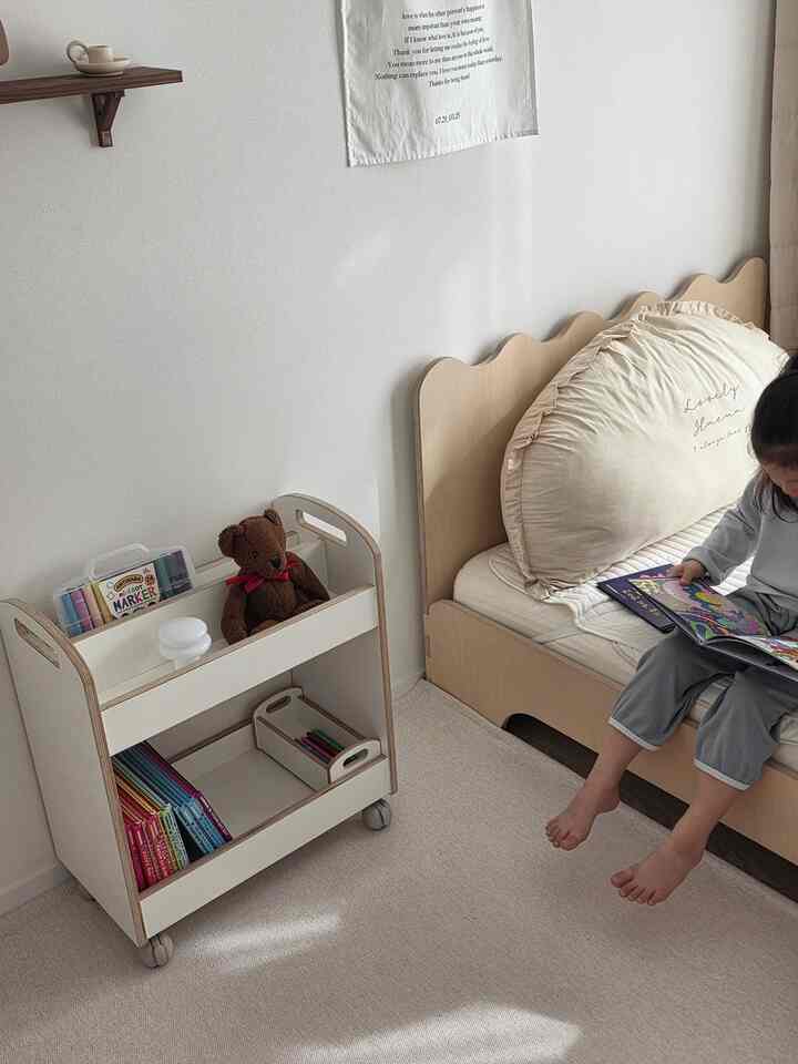 ベージュと木目調の子供部屋、低いベッドとブックカートが配置された清潔で温かみのあるインテリア