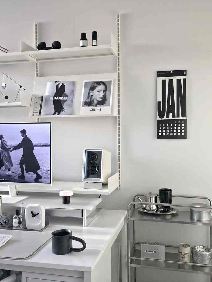Modern home office space in white and black tones featuring a desk, monitor, steel trolley, and coffee mugs with a clean atmosphere