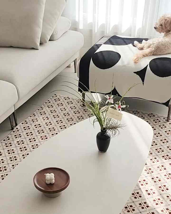 白とナチュラルカラーのリビングルーム、ベージュのソファとパターン柄のオットマン、のんびりとくつろぐ小さな犬がいるシンプルな空間