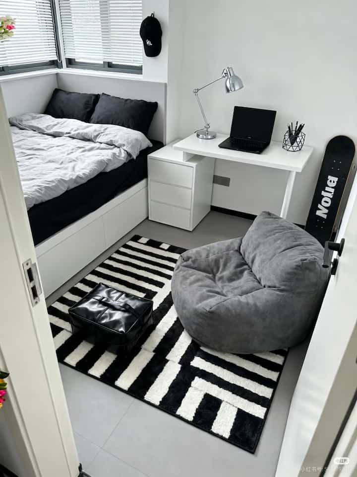Compact bedroom space with predominantly white and black tones, featuring a bed, desk with laptop, and a gray armchair in a modern setup