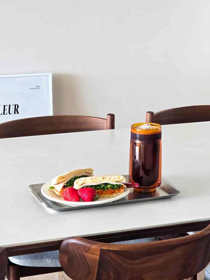 ベージュ色の壁と木製のダイニングチェアがあるキッチンスペース、白いテーブルの上にピタブレッドサンドイッチとアイスアメリカーノが置かれている印象的な空間