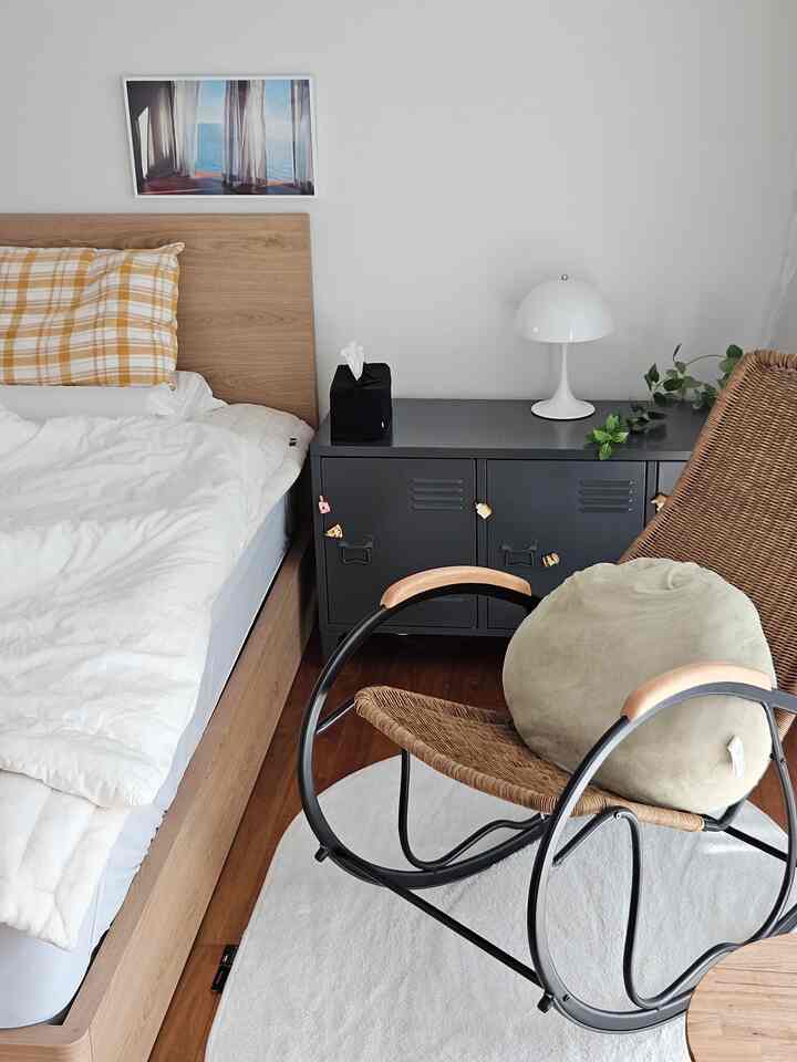 White and brown toned bedroom featuring a wooden bed, rattan armchair, nightstand and plants creating a cozy atmosphere