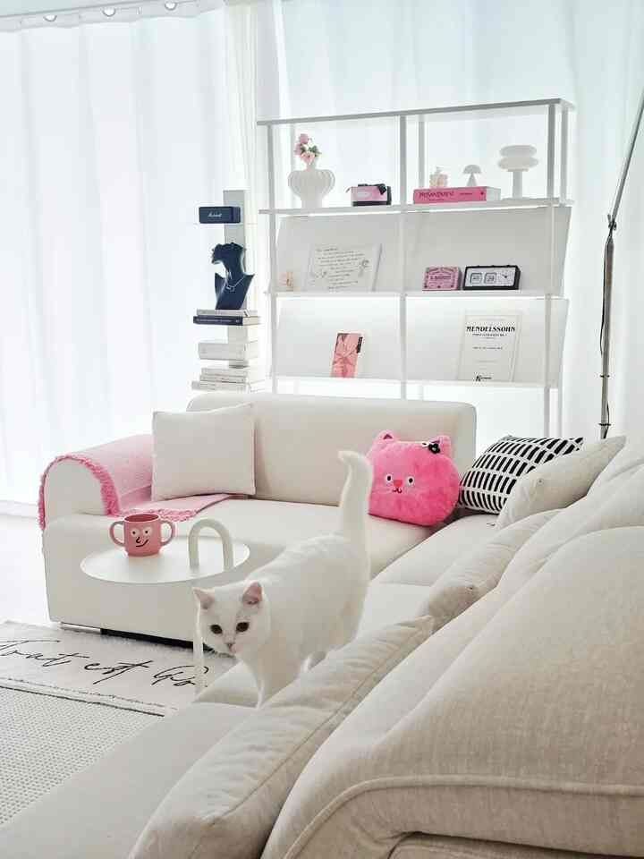 白とピンクのアクセントがあるリビング、広いソファと棚、猫がいるモダンで居心地の良い空間