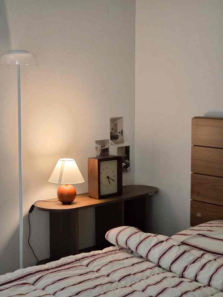 木目調とベージュの壁の寝室、ナイトテーブルにランプと時計が置かれた落ち着いたナチュラルな印象のインテリア