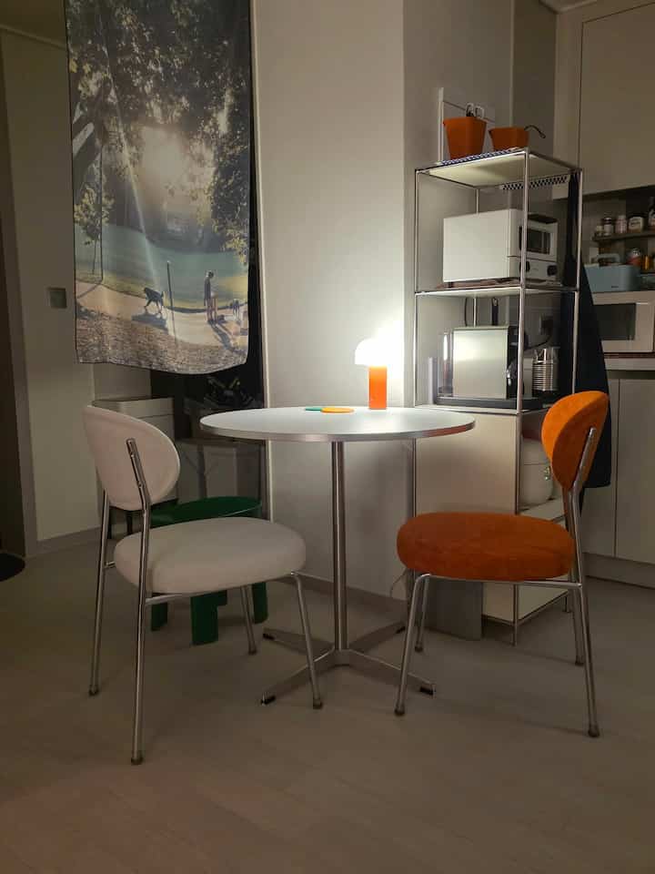 Compact kitchen space in white and orange tones featuring a round dining table with two chairs and a table lamp, creating a cozy simple interior.