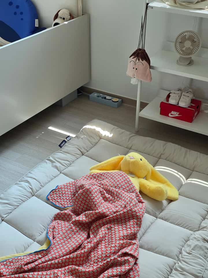 白い壁と木目調の床が調和する寝室、フロアマットレスとぬいぐるみが温かみのある雰囲気を醸し出しています。