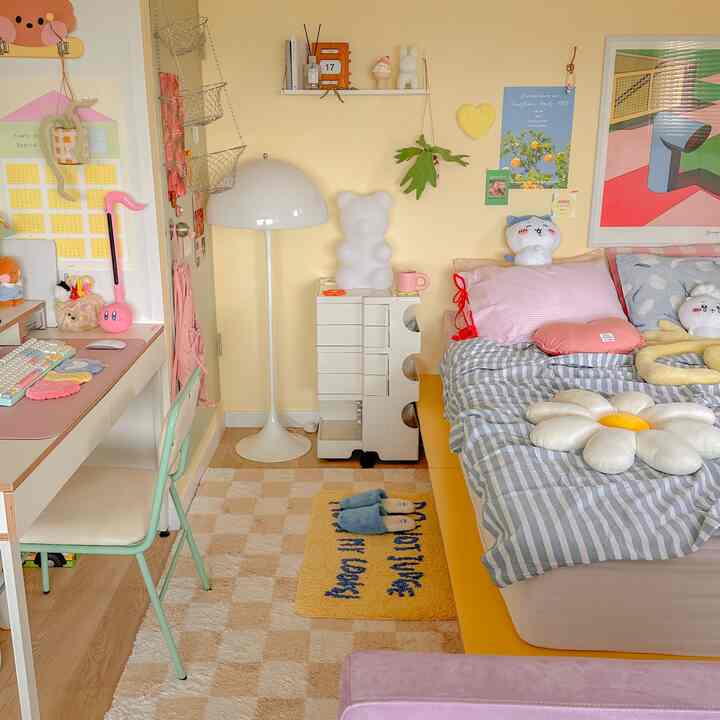 A cozy small bedroom in yellow and pink tones featuring a comfortable bed, desk, rug, and storage shelves