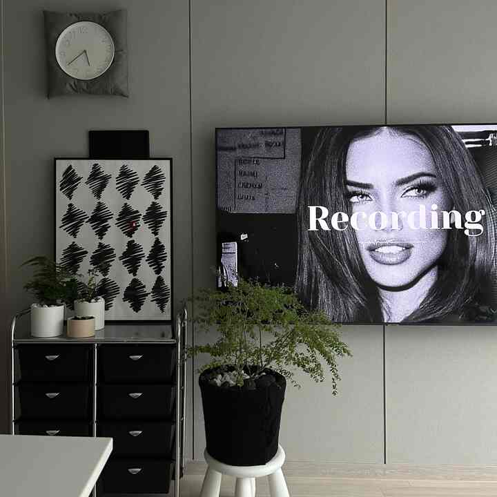 グレーの壁と白い天井のリビング、中央に植物と白いスツール、左に黒いチェストと時計、右に壁掛けテレビが配置されたモダンな空間