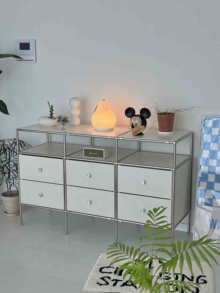 白を基調としたリビング、すっきりした金属製収納キャビネットと植物、暖かい照明が調和したミニマルな空間