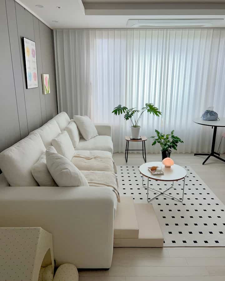 Bright white and natural toned living room featuring a beige sofa, round coffee table, and pet stairs with a dog house creating a cozy atmosphere