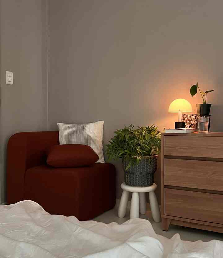 Gray-walled bedroom featuring wooden dresser, rust-colored single armchair, warm lamp lighting, and plants creating a cozy atmosphere