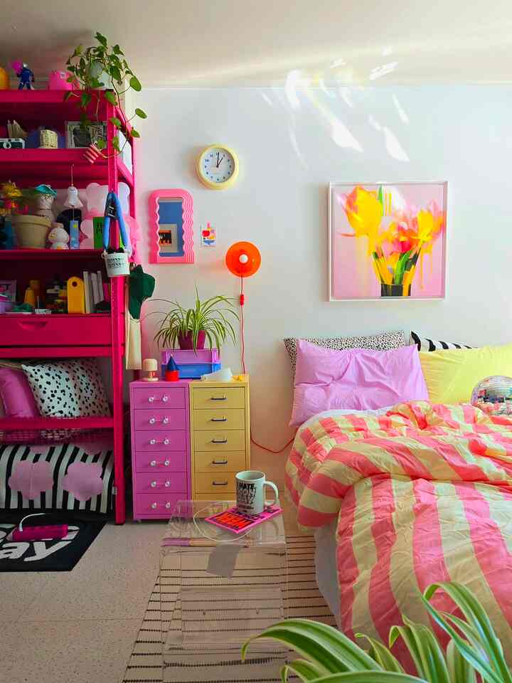 ピンクと黄色のトーンのワンルーム寝室、鮮やかな柄の寝具とチェスト、多彩な小物と植物が配置された明るく活気のあるインテリア
