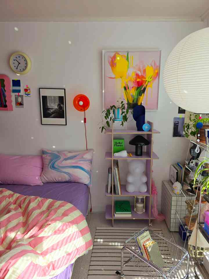 ピンクとパープルの色調の寝室、ストライプの布団と本棚、テーブルランプ、時計が配置された居心地の良い空間